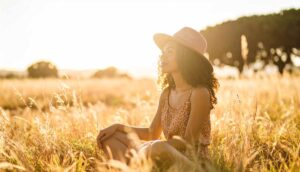 A young adult sitting cross-legged in a sunlit field, looking off into the distance with a thoughtful expression—evoking calm, curiosity, and quiet confidence.