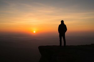 Silhouette of a person standing on a cliff at sunrise, looking out over a vast horizon.